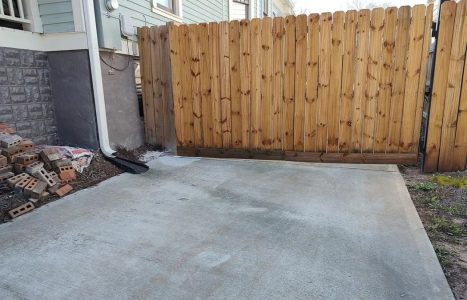 A wooden gate fully blocks a concrete driveway, preventing vehicle access; loose bricks and a bag of cement are stacked on the left side near the house.